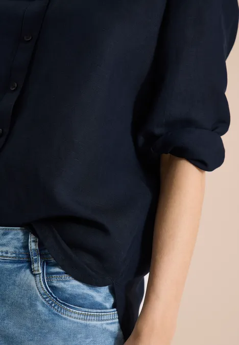 Teilansicht einer Person mit dunkelblauem Hemd und Jeans vor hellem Hintergrund.