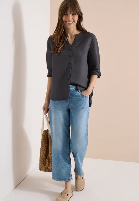 Eine Frau trägt eine dunkle Bluse mit 3/4-Ärmeln, weite blaue Jeans, beige Slipper und hält eine Korbtasche.