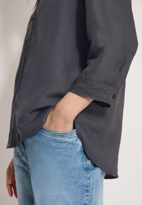 Schwarze Bluse über blaue Jeans, Hand lässig in der Hosentasche.