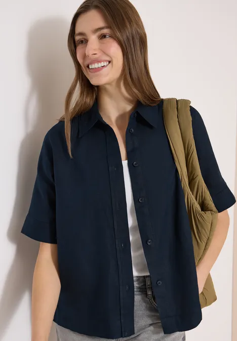 Schlanke Person mit schulterlangem Haar trägt dunkelblaues Kurzarmhemd über weißem Top, Jeans und olivgrüne Tasche.