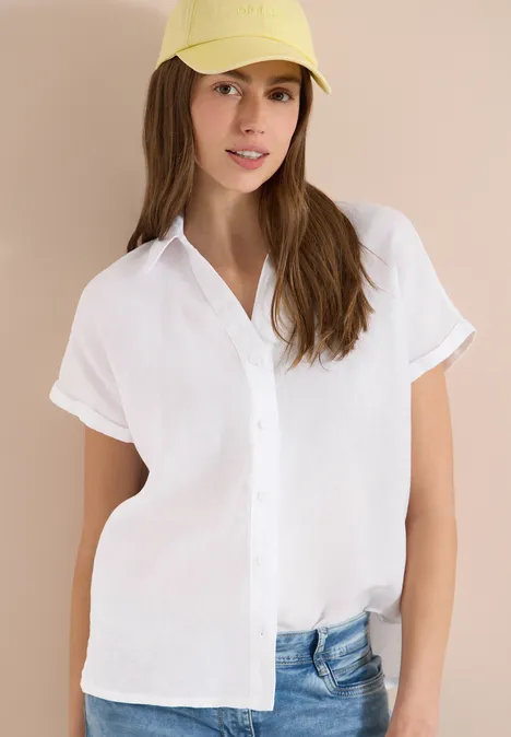 Jemand trägt eine weiße, kurzärmlige Bluse mit Knöpfen und eine blaue Jeanshose vor hellem Hintergrund.