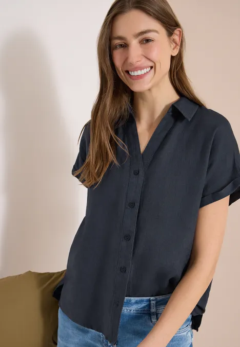 Frau in dunkelblauer Kurzarm-Bluse und Jeans vor hellem Hintergrund.