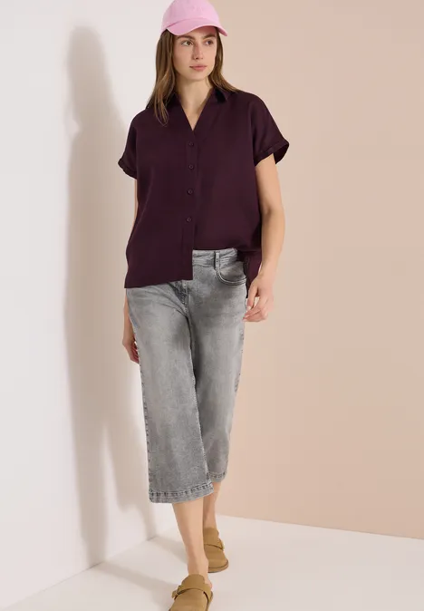 Eine Frau trägt ein kurzärmeliges, dunkelviolettes Hemd, graue Cropped-Jeans und beige Slipper vor hellem Hintergrund.