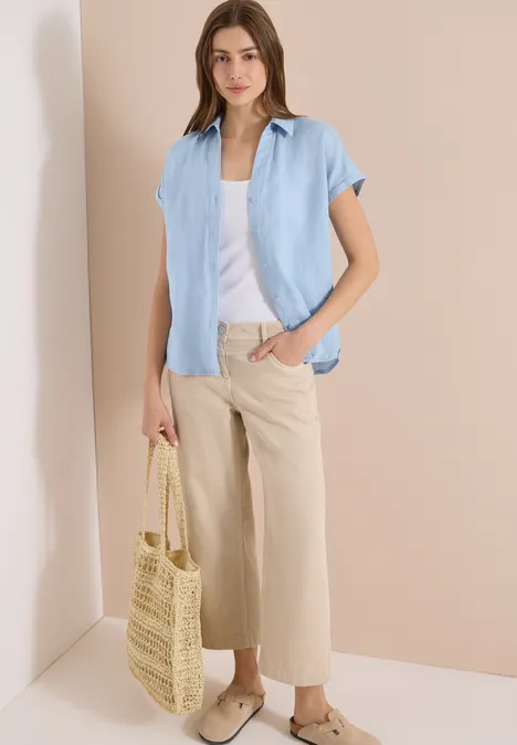 Frau in hellblauer Bluse, weißem Top, beigen Culottes und Strohtasche.