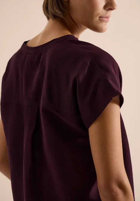 Rückansicht einer Frau in dunkelviolettem, fließendem Kurzarmshirt mit umgeschlagenen Ärmeln.