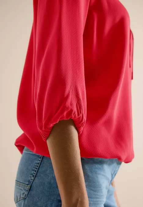 Seitliche Aufnahme einer Person in roter Bluse mit 3/4-Ärmeln und blauen Jeans.