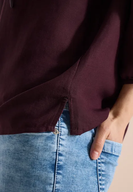 Detailaufnahme einer Person mit dunkelviolettem Oberteil und hellblauen Jeans, die Hand in der Hosentasche.