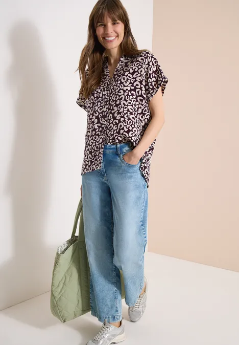 Eine Frau trägt ein geblümtes Oberteil, weite Jeans, weiße Sneaker und eine grüne Stepp-Tasche.