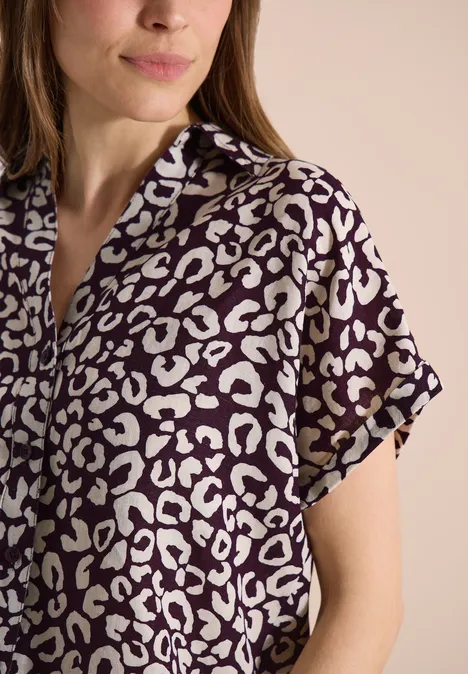 Frau in kurzärmligem, lila-weißem Hemd mit Leopardenmuster vor neutralem Hintergrund.