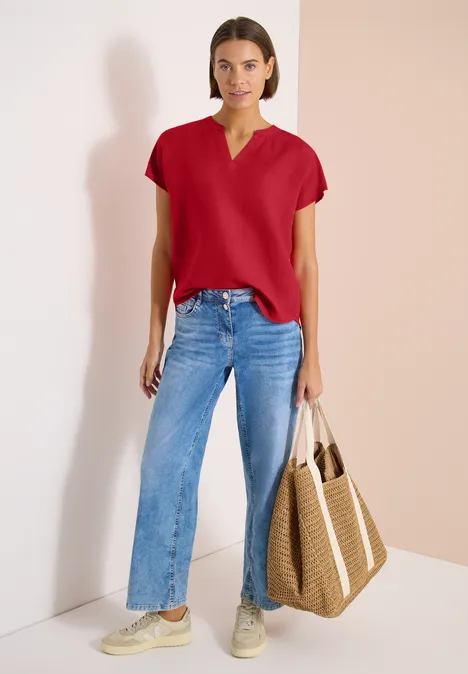 Frau vor hellem Hintergrund in rotem Shirt, blauen Jeans, Sneakern und geflochtener Strohtasche.