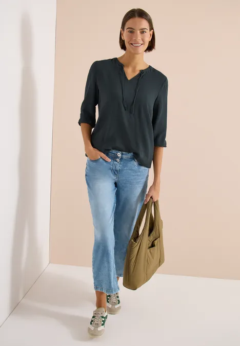 Frau steht vor hellem Hintergrund, trägt dunkles Oberteil, blaue Jeans, Sneaker und eine grüne Umhängetasche.