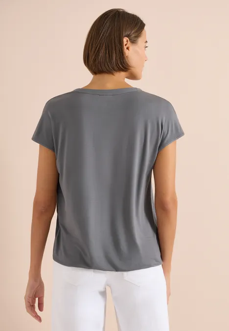 Rückansicht einer Person in grauem T-Shirt und weißer Hose vor neutralem Hintergrund.