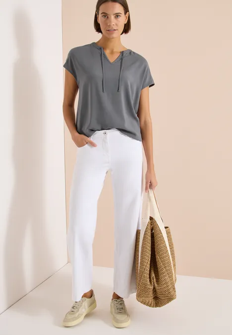 Frau trägt graues Shirt, weiße Hose und Strohtasche vor beiger Wand.