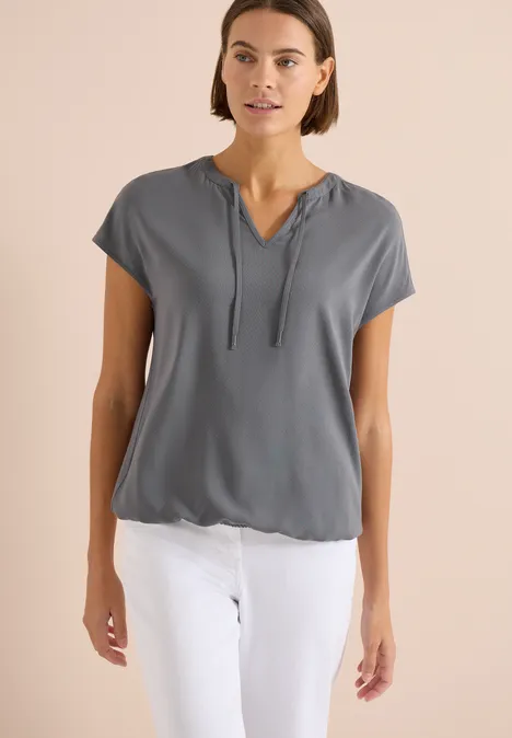 Frau in grauer Kurzarm-Kapuzenbluse mit V-Ausschnitt und weißen Hosen.