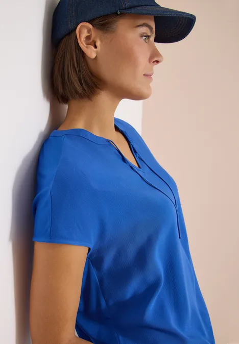 Frau mit blauem Kurzarmtop und dunkelblauem Basecap lehnt an heller Wand.