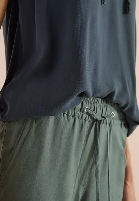 Eine Person trägt ein dunkelblaues ärmelloses Oberteil und olivgrüne Hosen mit Kordelzug.