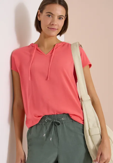Frau lehnt an Wand, trägt pinkes Kurzarm-Kapuzenshirt, grüne Hose und beigen Stoffbeutel.