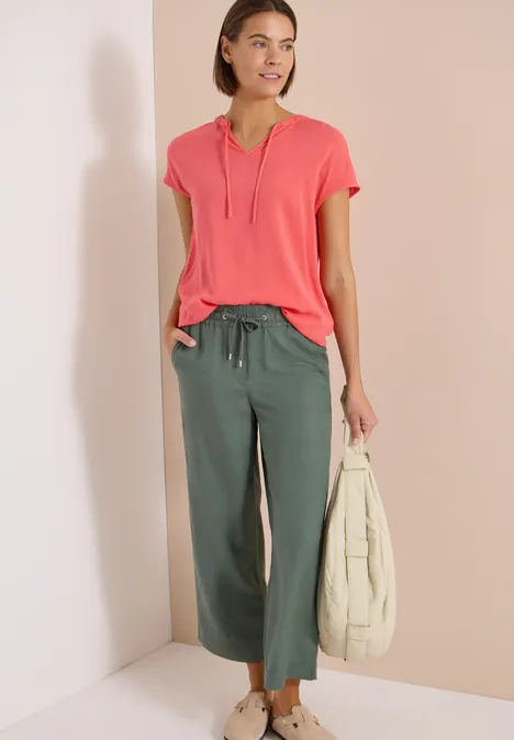 Frau trägt pinkes T-Shirt mit V-Ausschnitt, grüne Culotte-Hose und hält eine weiße Tasche.