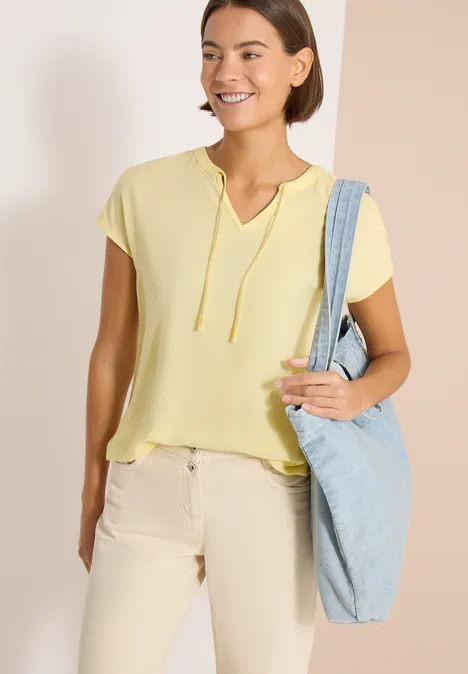 Vrouw met geel T-shirt en korte mouwen draagt een lichtblauwe schoudertas, neutrale achtergrond.