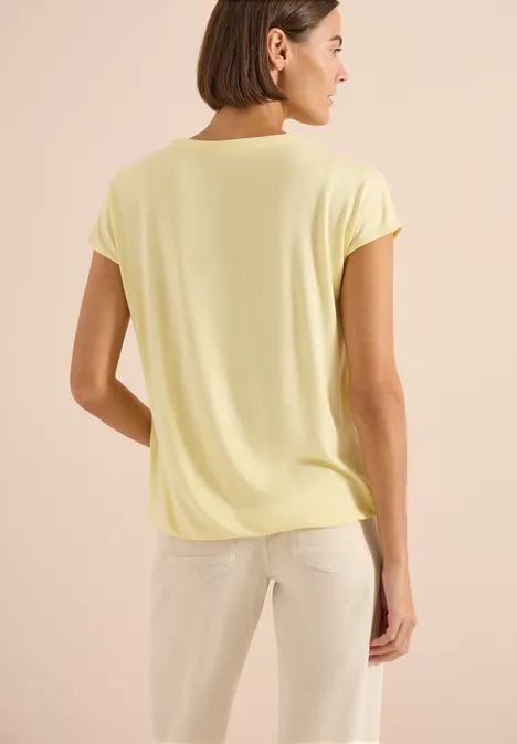 Persoon van achteren met lichtgeel shirt en lichte broek voor een beige achtergrond.