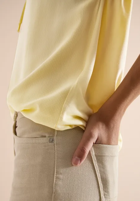 Iemand draagt een lichtgele blouse en heeft de hand in de zak van een beige broek.