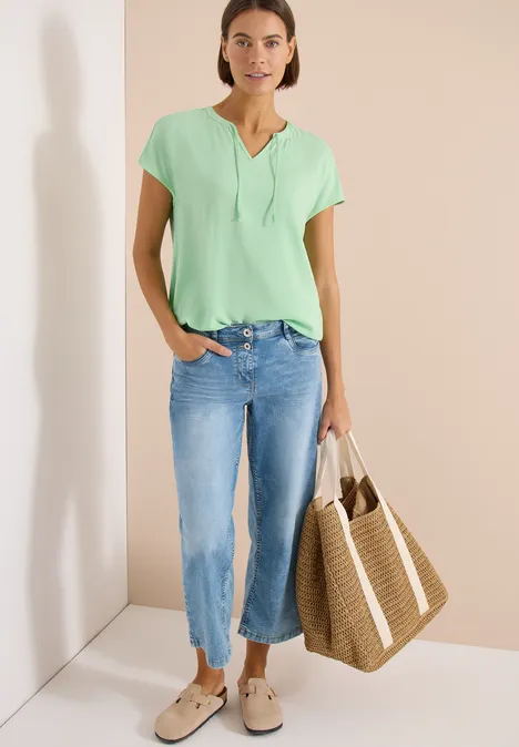 Vrouw in groen t-shirt en blauwe jeans met grote strooien tas, staand tegen een lichte muur.