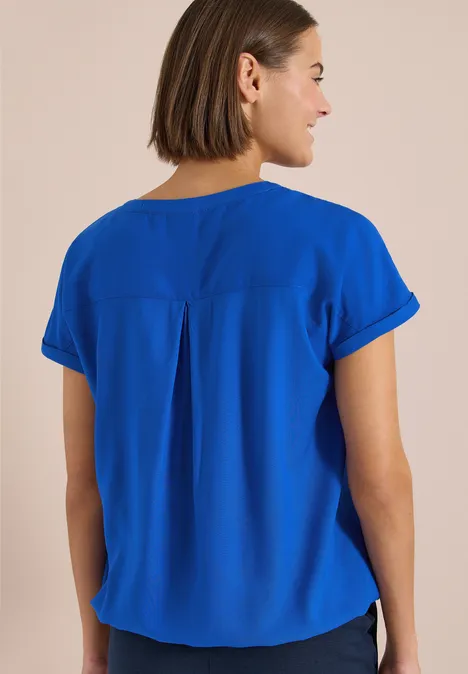 Rückenansicht einer Frau mit kurzem braunen Haar in leuchtend blauer Bluse vor hellem Hintergrund.