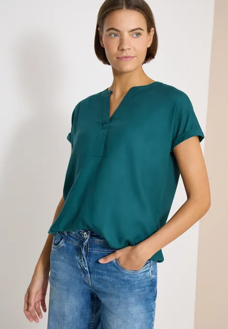 Vrouw draagt donkergroen kort mouw blouse met knoopsluiting en blauwe spijkerbroek.