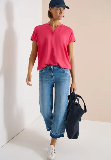 Persoon in roze shirt, blauwe jeans en witte sneakers, met een donkerblauwe tas.