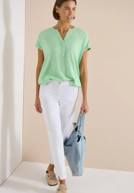 Vrouw in mintgroene blouse, witte broek en beige instappers, draagt een lichtblauwe tas.