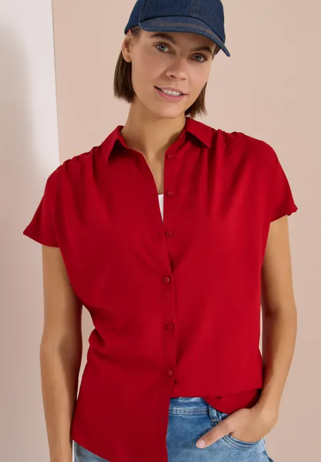 Personne porte une chemise rouge à manches courtes et un jean bleu, une main dans la poche, casquette noire.