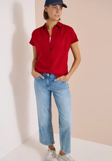 Femme en polo rouge et jean bleu, mains dans les poches, posant détendue dans un coin sur fond clair.