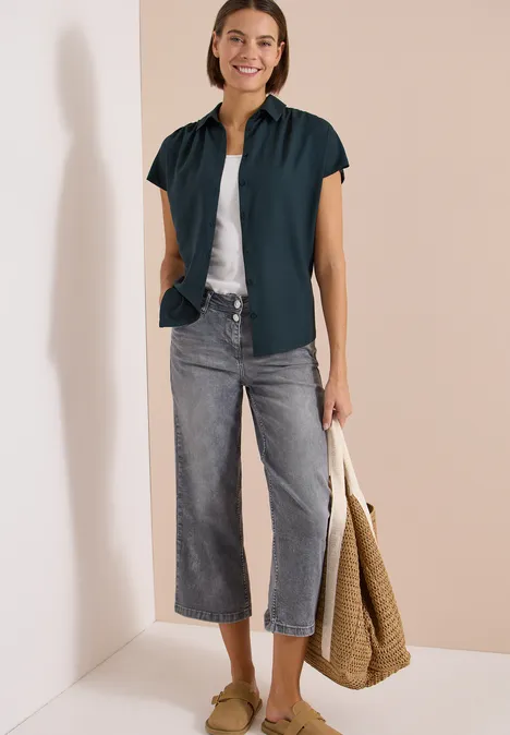 Frau in dunklem kurzärmligen Hemd, weißem Top, grauen Culotte-Jeans, Sandalen und Korbtasche.