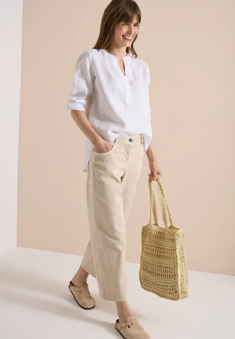 Frau in weißer Leinenbluse, beigen Cropped-Hosen, geflochtenen Espadrilles und Strohtasche vor hellem Hintergrund.