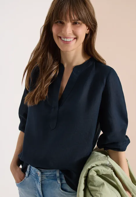 Frau in dunkelblauer Bluse und Jeans, hält eine grüne Jacke in der Hand.
