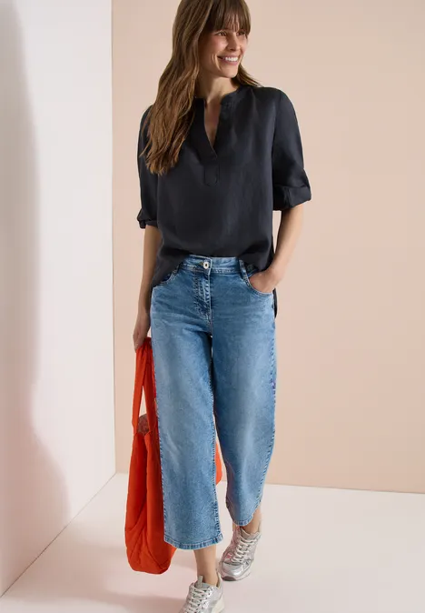 Frau trägt schwarze Bluse, weite Jeans, weiße Sneaker und eine orangefarbene Tasche vor neutralem Hintergrund.