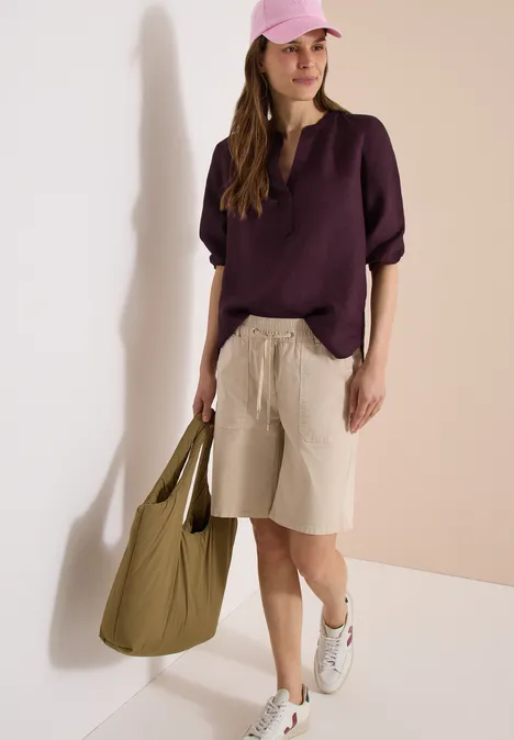 Frau trägt lila Bluse, beige Bermudashorts, weiße Sneaker und große grüne Tasche vor neutralem Hintergrund.