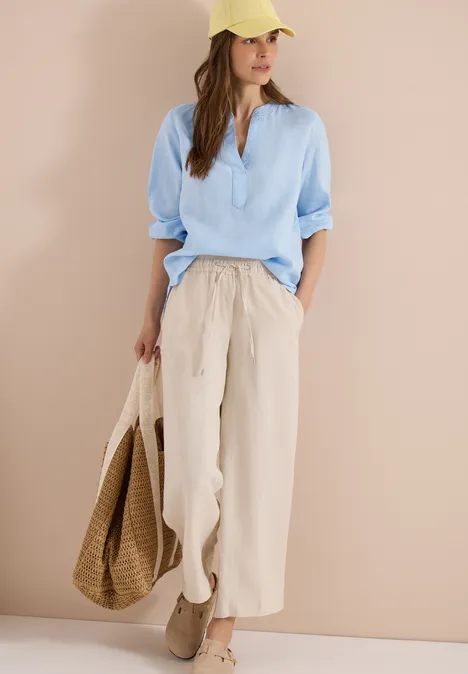 Eine Frau trägt weite Leinenhose in Beige, hellblaues Hemd, gelbe Mütze, hält einen Korbbeutel