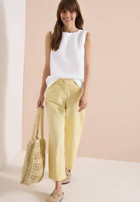Frau in weißem ärmellosem Top, gelben Hosen und geflochtener Tasche, vor hellem Hintergrund