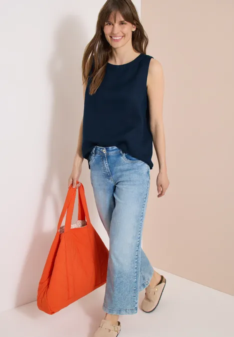 Femme en haut bleu sans manches, jean ample et chaussures beige, portant un grand sac rouge.