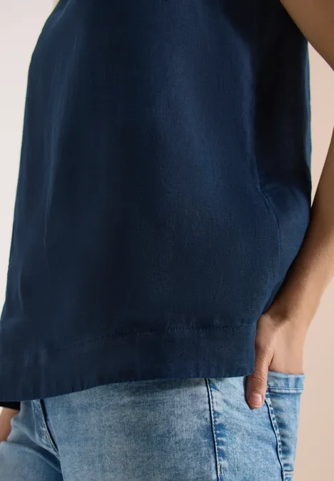 Une personne porte un haut sans manches bleu foncé et un jean clair, main dans la poche.