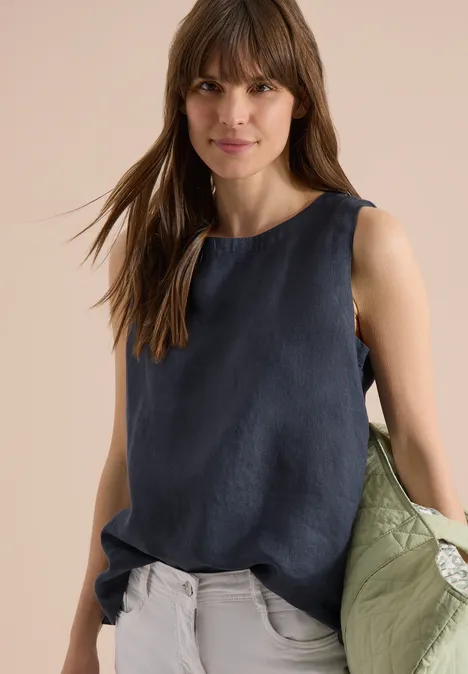 Femme en top sans manches bleu marine, pantalon clair et manteau vert tenu à la main.