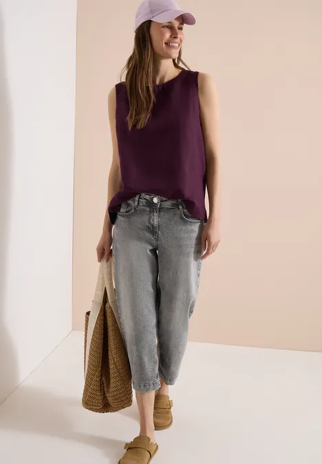 Person mit lila Top, weiten grauen Jeans, Cap, Basttasche und Clogs