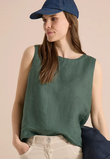 Eine Frau trägt ein ärmelloses, grünes Top, beige Hosen und eine blaue Mütze, Hände in den Hosentaschen.