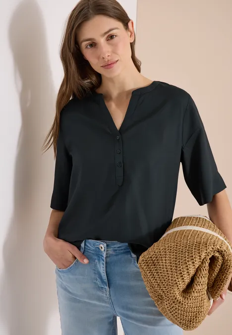 Frau in schwarzer Bluse und blauen Jeans hält eine beige Strickjacke in der Hand.