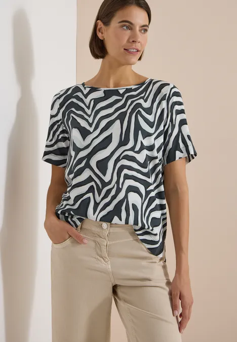 Frau trägt ein kurzärmeliges Zebra-Muster-Oberteil und beige Hose vor neutralem Hintergrund.