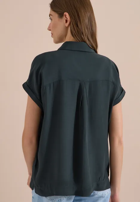 Chemisier à manches courtes uni palm breeze green Vue de dos d'une femme portant une blouse colletée à manches courtes vert foncé devant un fond beige uni.