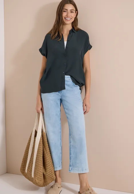 Chemisier à manches courtes uni palm breeze green Femme sur fond beige, chemise noire à manches courtes, jean large bleu clair et grand sac en paille.