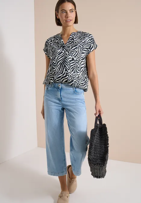 Frau mit zebragemustertem Oberteil, weiten Jeans, Basttasche und Espadrilles.