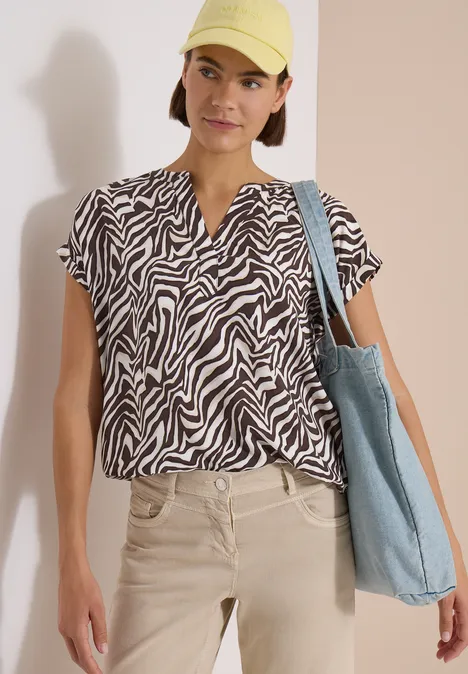 Femme en chemisier zèbre, pantalon beige, casquette jaune et sac en toile bleu clair devant un fond neutre.